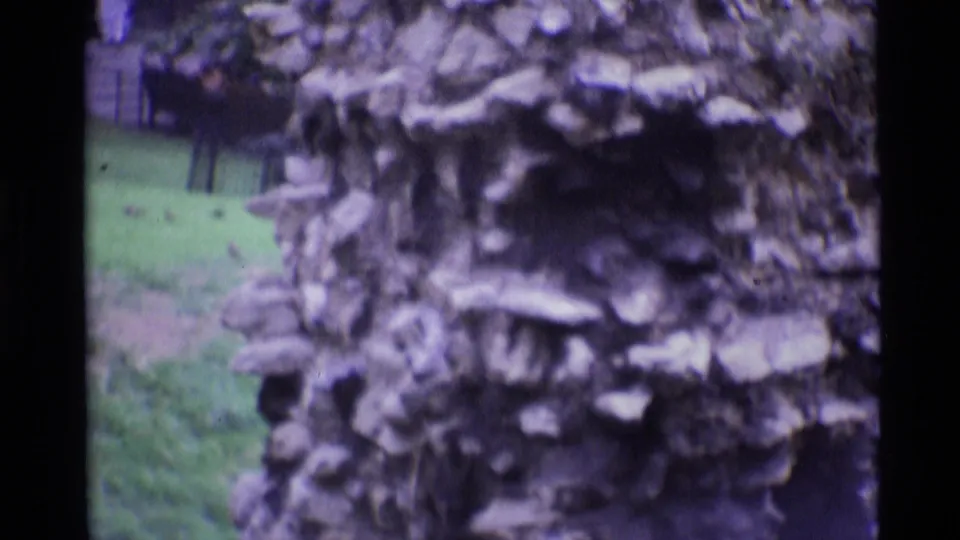 1969 London home-movie frame showing weathered stonework, grass, and nearby greenery in a garden-like exterior setting.