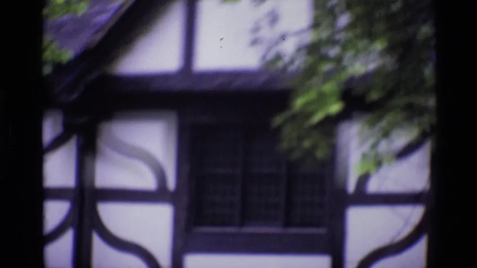 1969 London 8mm frame of a half-timbered house facade, with a dark window, white wall panels, and overhanging greenery.