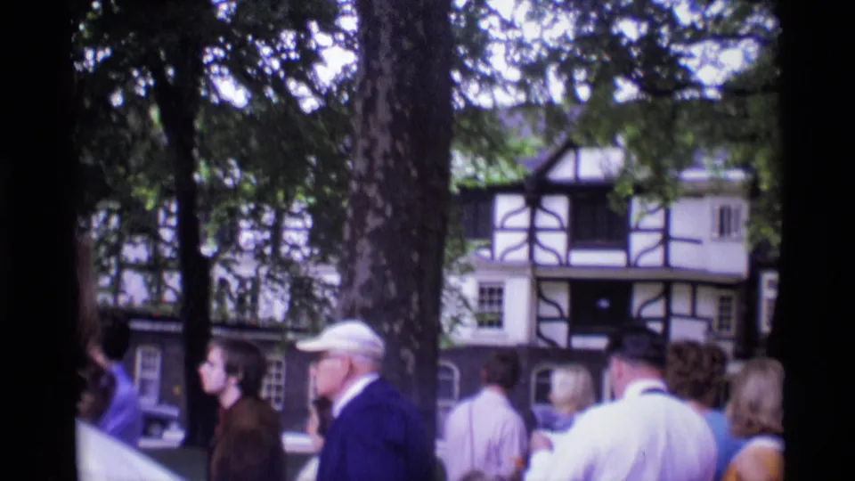 A visitor-shot 1969 London frame shows adults near large trees and a half-timbered building, including a man in a white cap.