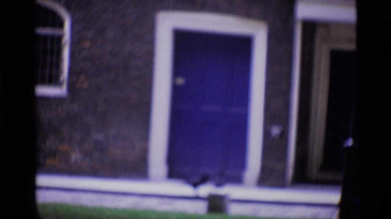 1969 London home-movie frame showing a blue front door, brick wall, white trim, lawn, and a small bird near the entrance.