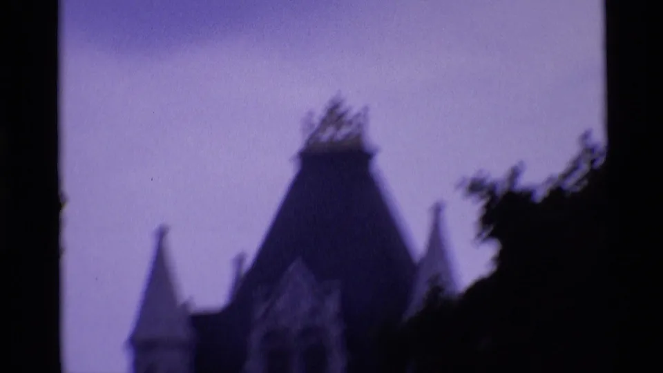 Soft 1969 London home-movie view of a steep tower roof and smaller roof peaks silhouetted against a purple-tinted sky.