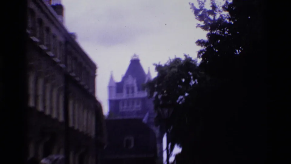 1969 London 8mm street-level view with an older building at left, trees at right, and a distant steep-roofed stone structure.