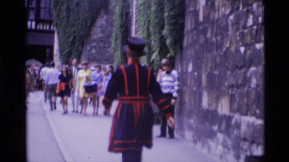 A Yeoman Warder walks through a narrow London landmark passage with tourists following in the background.