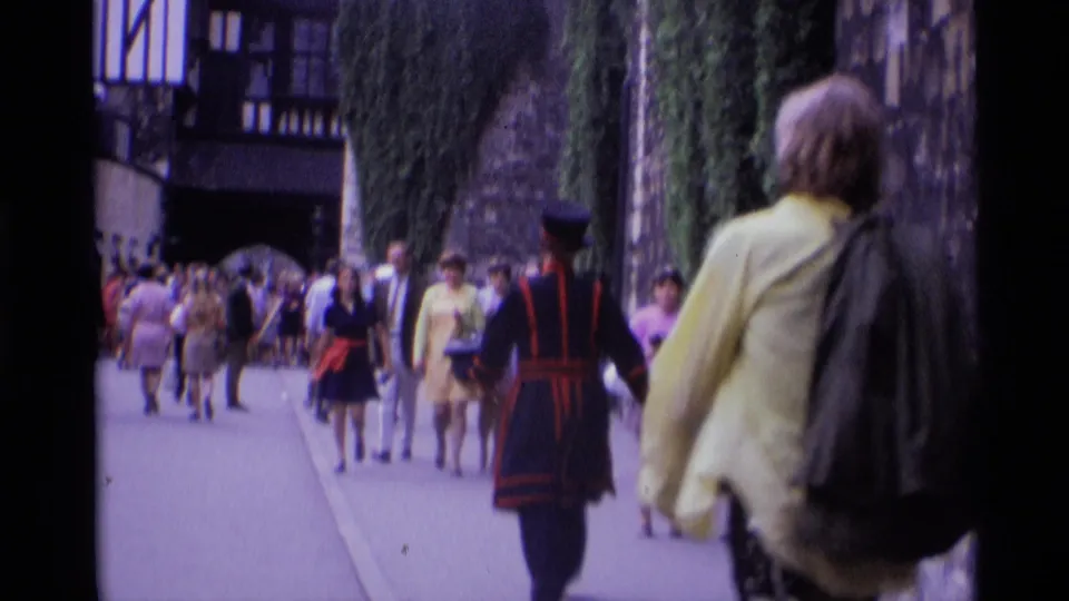 Tourists in 1960s dress pass a Yeoman Warder along an ivy-lined walkway at a London historic site.