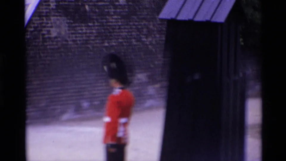 1969 London home-movie frame of a Royal Guard in red uniform standing beside a stone wall near a dark sentry box or post.