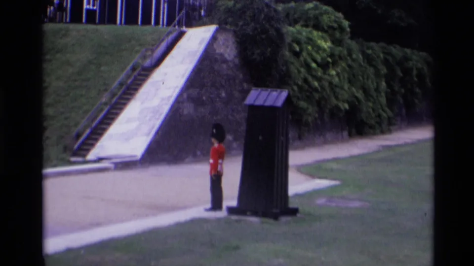 1969 London 8mm frame showing a Royal Guard at attention near a dark sentry box, with grass, steps, shrubs, and paths behind.