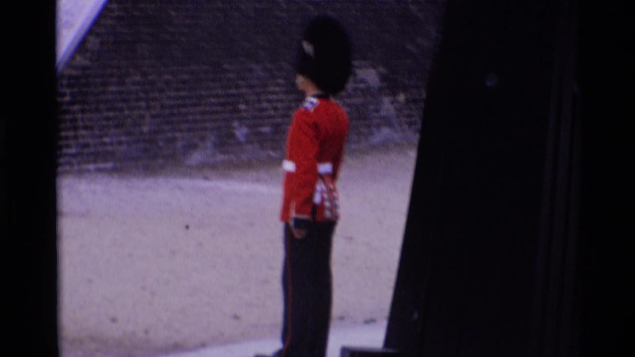1969 London home-movie frame showing a Royal Guard in red tunic and tall black hat standing at attention near stonework.