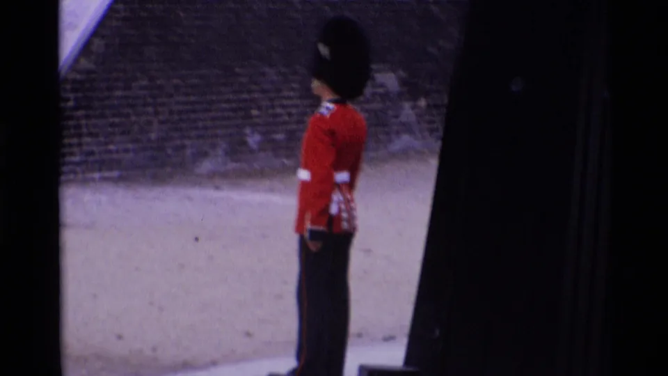 1969 London home-movie frame showing a Royal Guard in red tunic and tall black hat standing at attention near stonework.