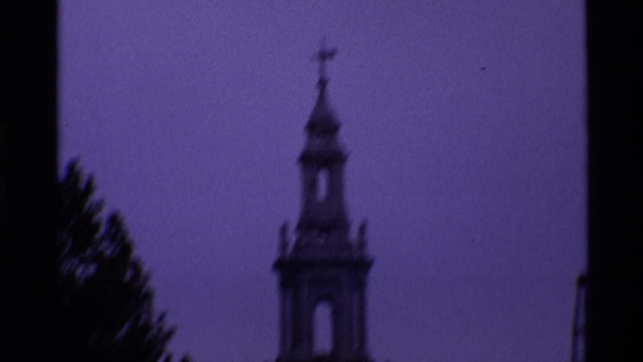 Purple-tinted 1969 London frame with a church spire and cross seen in soft focus beside dark trees.