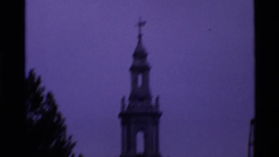Purple-tinted 1969 London frame with a church spire and cross seen in soft focus beside dark trees.