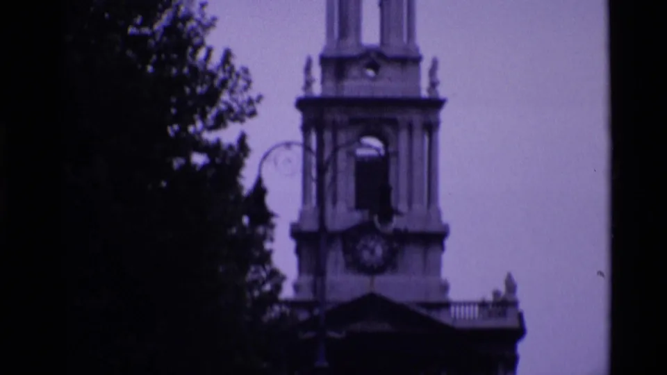 1969 London 8mm view of a church tower with a clock face, bell opening, and trees in the foreground.
