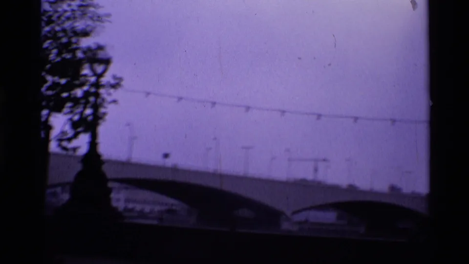 Soft 8mm London frame showing bridge arches, overhead wires, and a silhouetted tree beside the river.