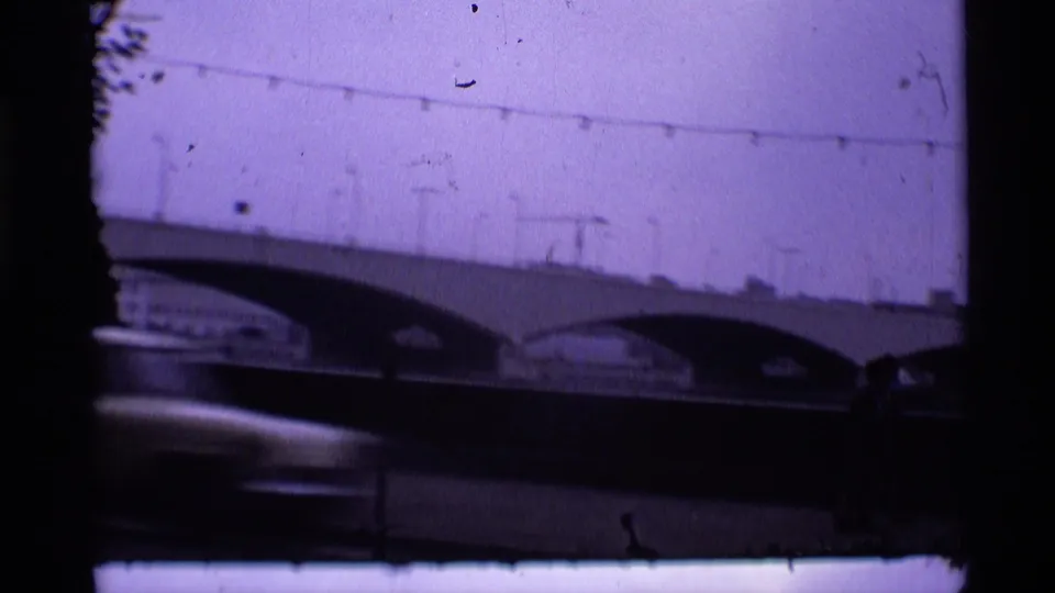1969 London 8mm frame showing the bridge, lamp line, dark foreground traffic area, and small indistinct people near the right edge.