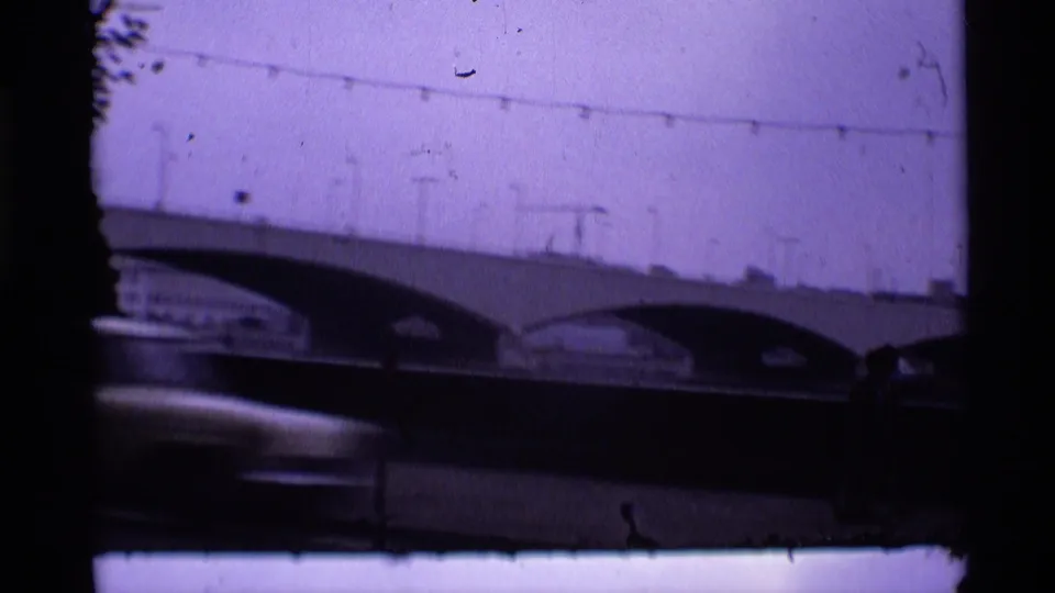 1969 London home-movie frame with the bridge spanning the scene, overhead wires, film scratches, and a passing car blur.