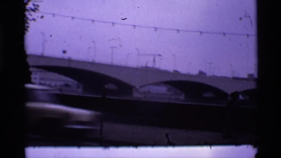 1969 London 8mm still with bridge arches, a blurred foreground car, overhead lines, and visible gate weave and edge shadows.