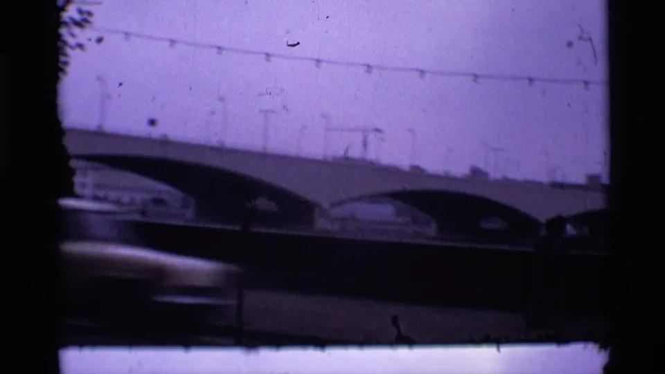 1969 London home-movie frame showing the arched bridge, overhead wires, purple color cast, and a vehicle blurred along the bottom edge.