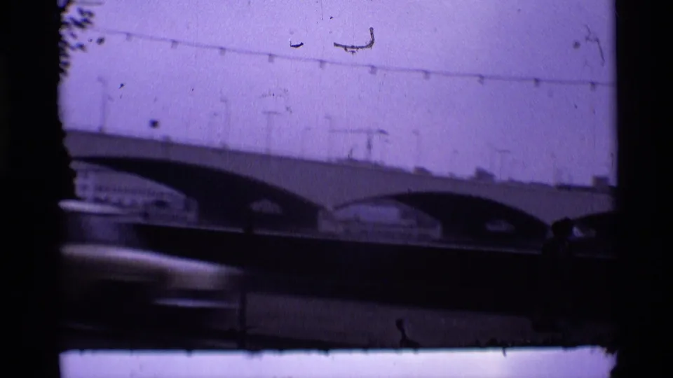 Purple-shifted 1969 London home-movie frame of an arched bridge spanning water, with small figures near the lower right.
