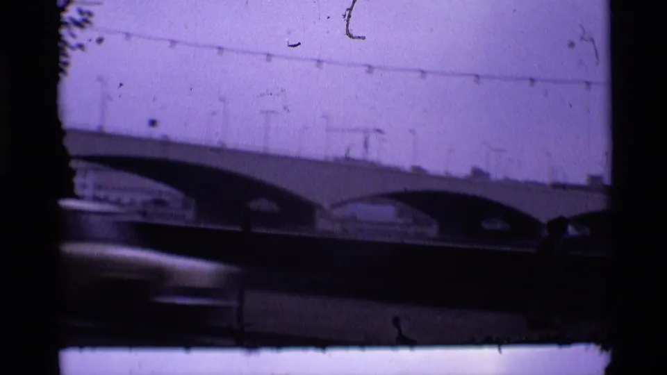 1969 London home-movie frame showing bridge arches, traffic movement below, pale sky, and visible 8mm film wear.