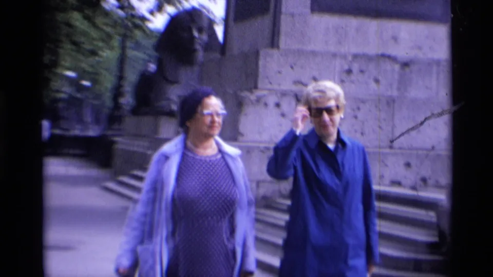 Two women in coats walk toward the camera in 1969 London, passing a stone monument base with a lion statue and steps behind them.