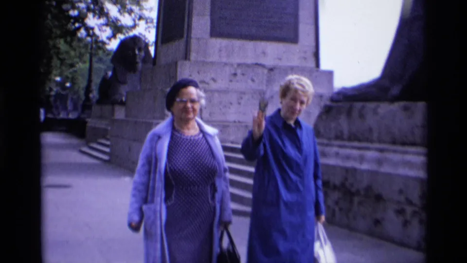 1969 London home-movie frame of two women with bags beside a stone monument, with one woman raising her hand toward the camera.