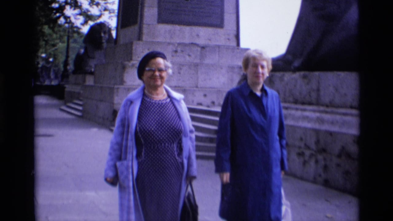 Two adult women in dresses and coats walk toward the camera on a London pavement beside a large stone monument.
