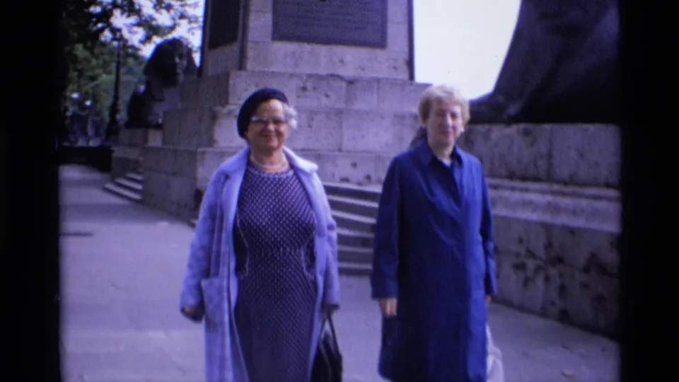Two adult women in dresses and coats walk toward the camera on a London pavement beside a large stone monument.