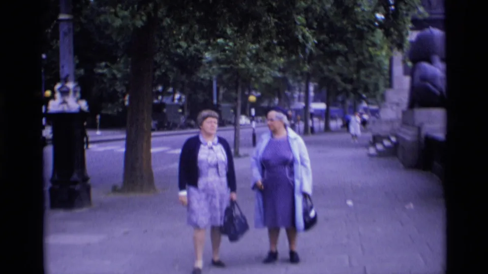 Two adult women walk side by side on a London sidewalk with trees, lamp posts, parked cars, and a monument edge nearby.