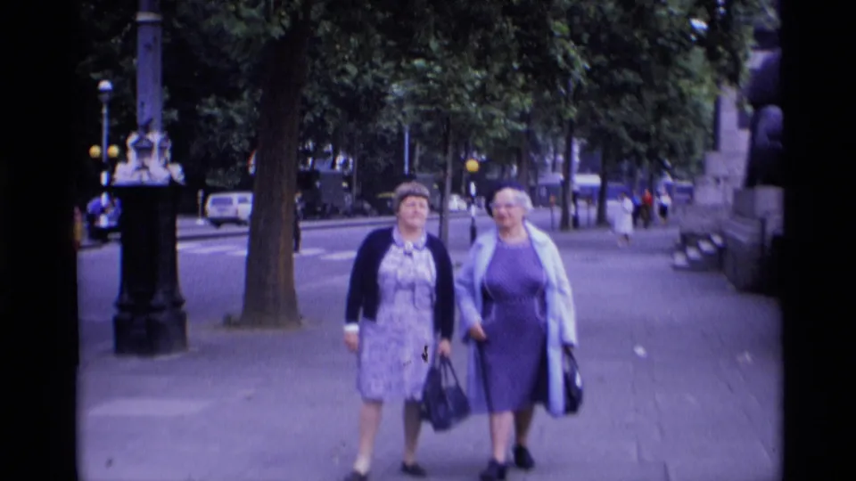 Two women in 1960s-style dresses, hats, and coats walk toward the camera along a London pavement bordered by trees and street lamps.