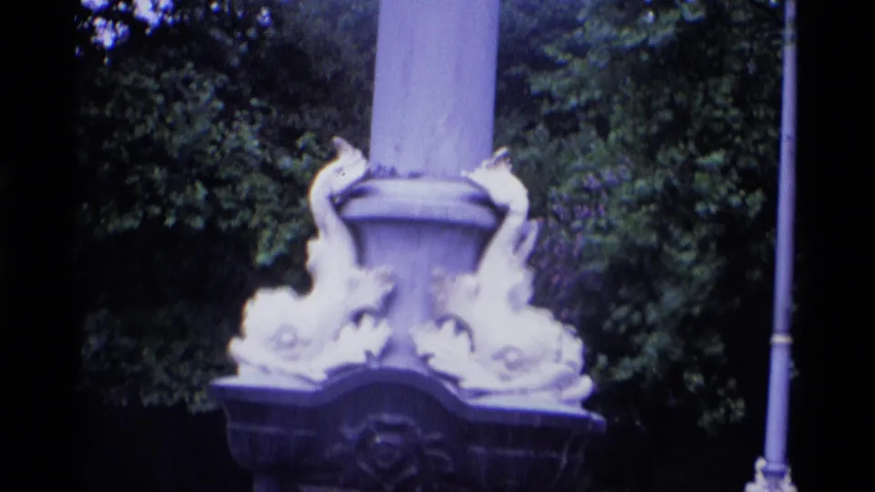 1969 London 8mm frame showing an ornate lamp base near dense trees and a roadside setting.