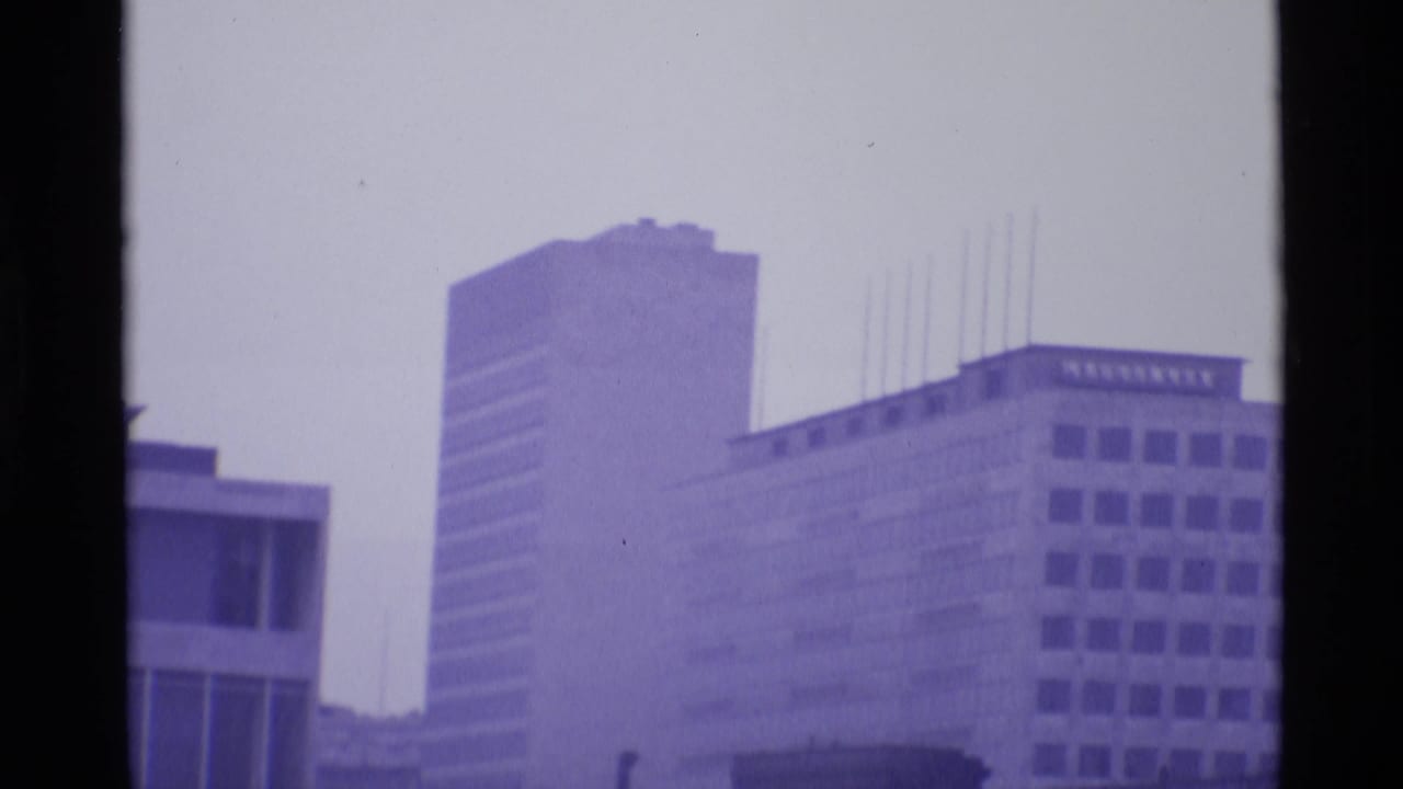 1969 London home-movie frame showing high-rise office blocks and modernist buildings in a purple-tinted haze.