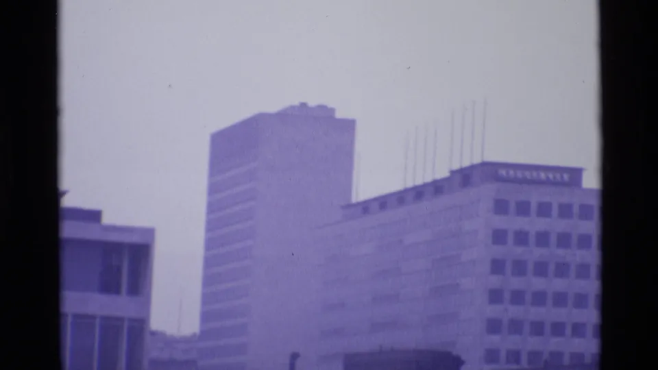 1969 London home-movie frame showing high-rise office blocks and modernist buildings in a purple-tinted haze.