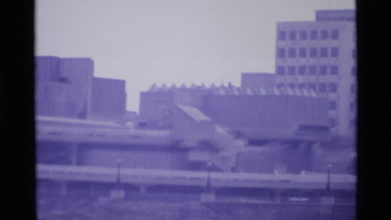 1969 London home-movie frame showing blurred modern urban buildings against a pale sky with strong purple film tint.