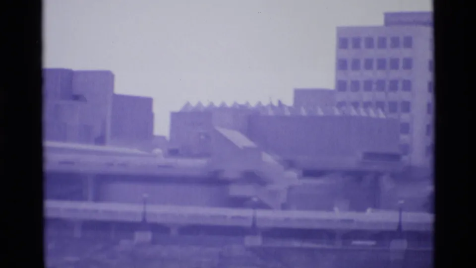 1969 London home-movie frame showing blurred modern urban buildings against a pale sky with strong purple film tint.