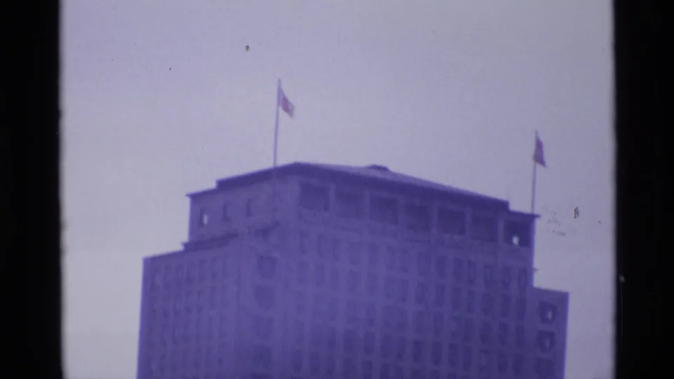 1969 London frame of a tall, windowed urban building with flagpoles on the roof, seen through soft purple-tinted 8mm footage.