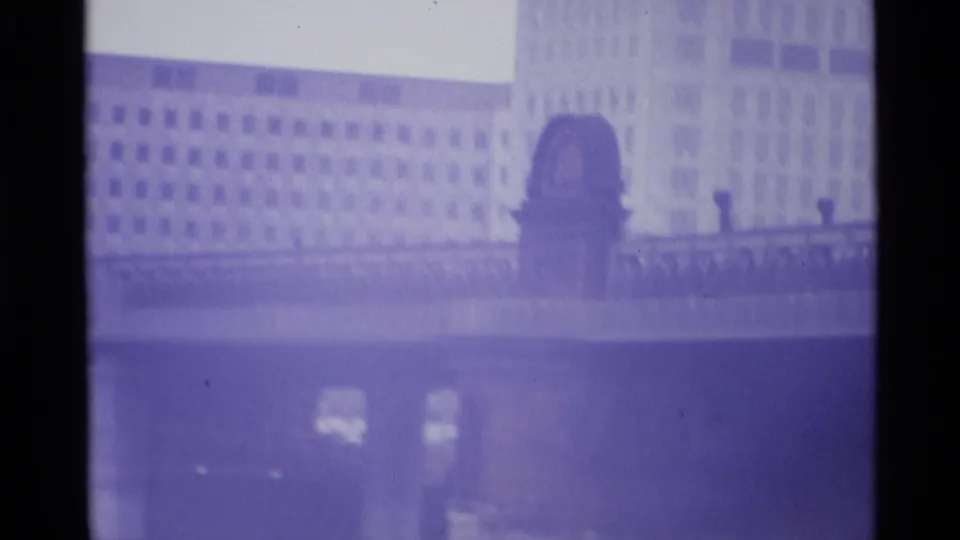 1969 London frame showing a broad modern facade with an arched central feature and pale high-rise forms in the background.