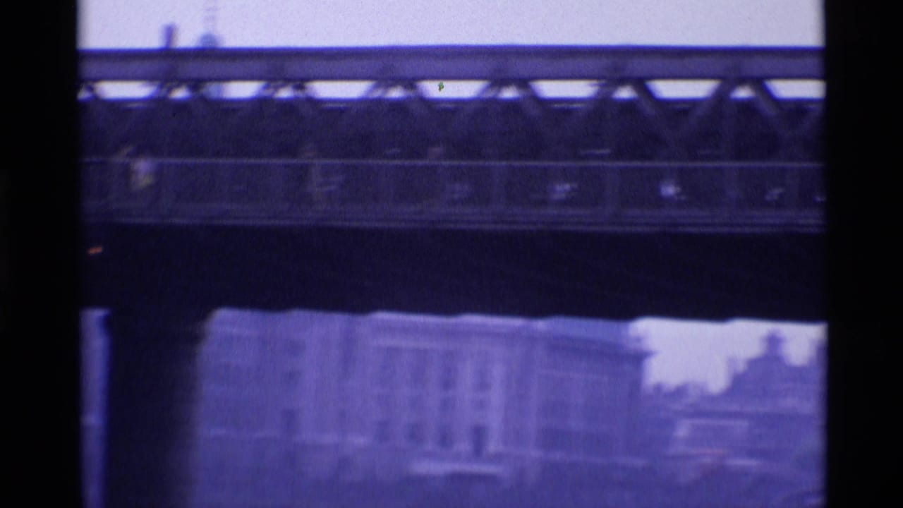 1969 London home-movie frame showing a bridge span through dark framing, with soft-focus buildings and riverfront shapes beyond.