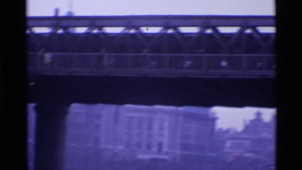 1969 London home-movie frame showing a bridge span through dark framing, with soft-focus buildings and riverfront shapes beyond.