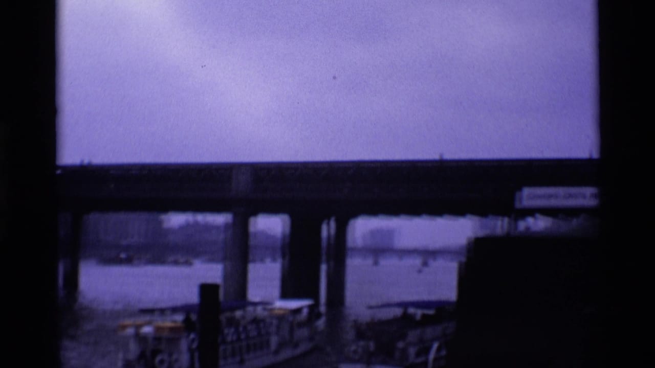 London riverfront view beneath a bridge, showing pier railings, water, distant boats, and dark structural framing.