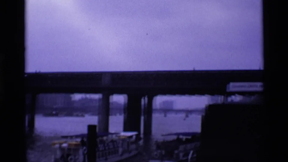 London riverfront view beneath a bridge, showing pier railings, water, distant boats, and dark structural framing.