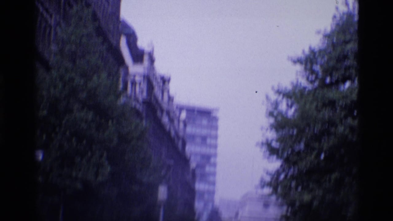 1969 London 8mm frame showing a blurred tree-lined street with older facades and a modern block beyond.