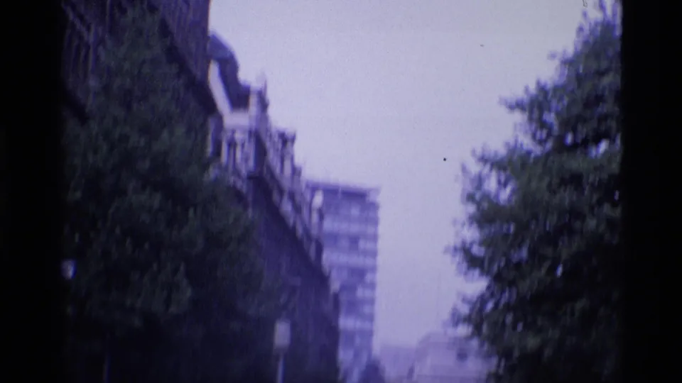 1969 London 8mm frame showing a blurred tree-lined street with older facades and a modern block beyond.