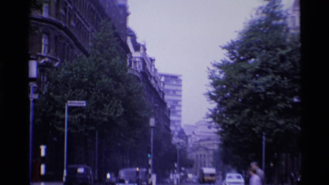 1969 London street view with classic cars, traffic lights, trees, and tall urban buildings along the road.