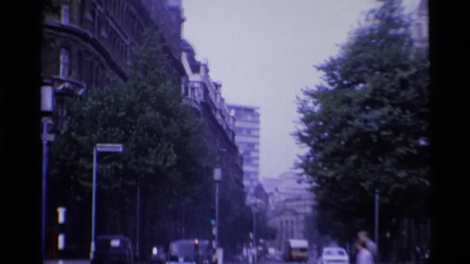 1969 London street view with classic cars, traffic lights, trees, and tall urban buildings along the road.