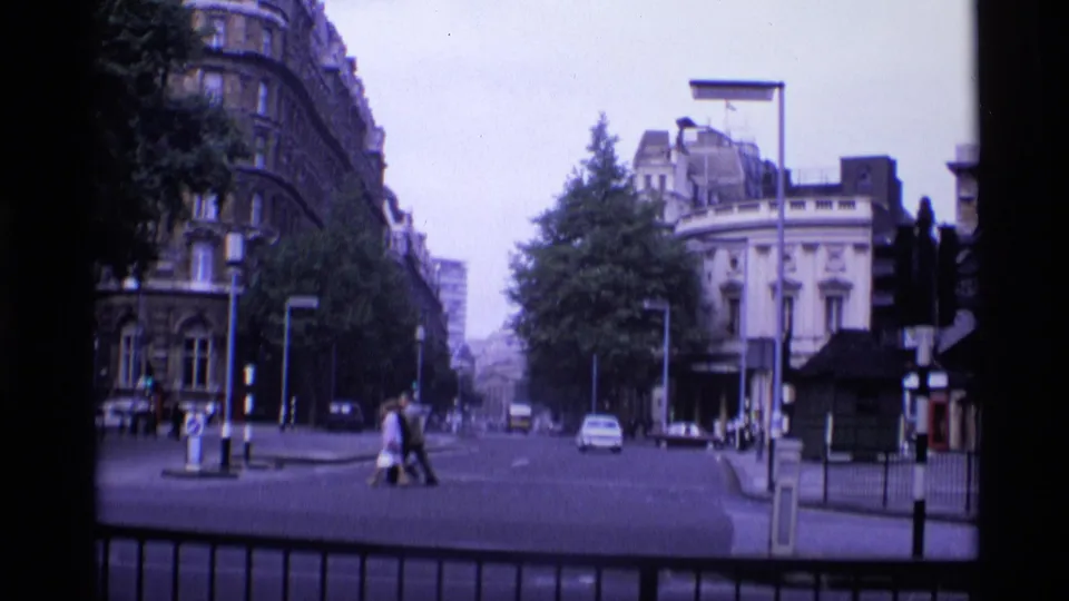 1969 London frame with a couple crossing the street beside classic cars, traffic lights, trees, and ornate architecture.