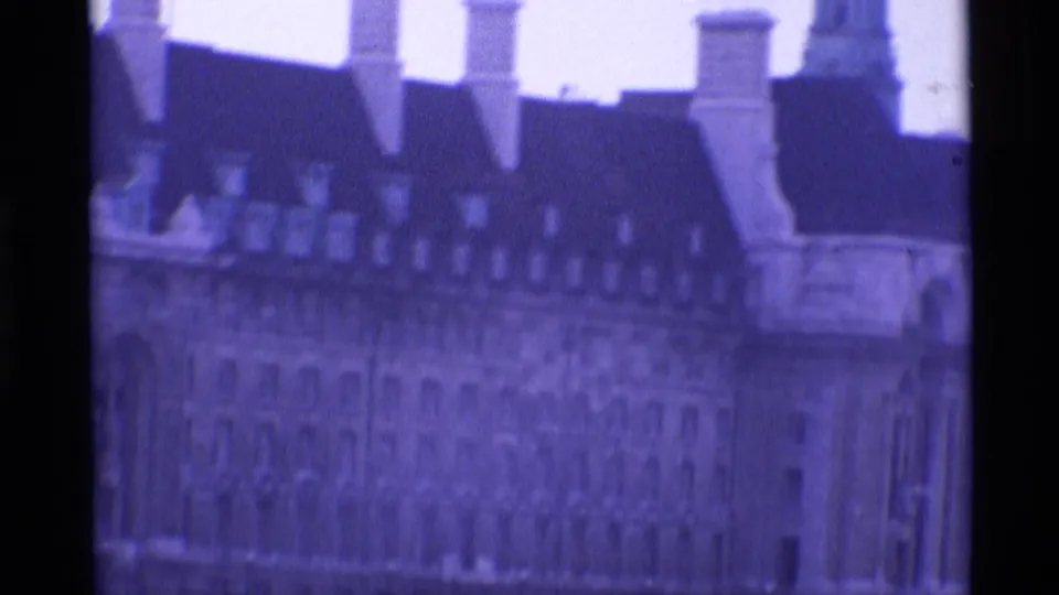 1969 London frame of a grand stone facade with mansard-style roof, many windows and tall chimneys in soft purple-tinted 8mm footage.