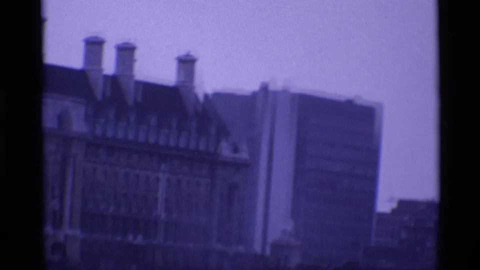 1969 London frame showing ornate roof peaks and chimneys in front of a modern high-rise building, recorded on purple-tinted 8mm film.