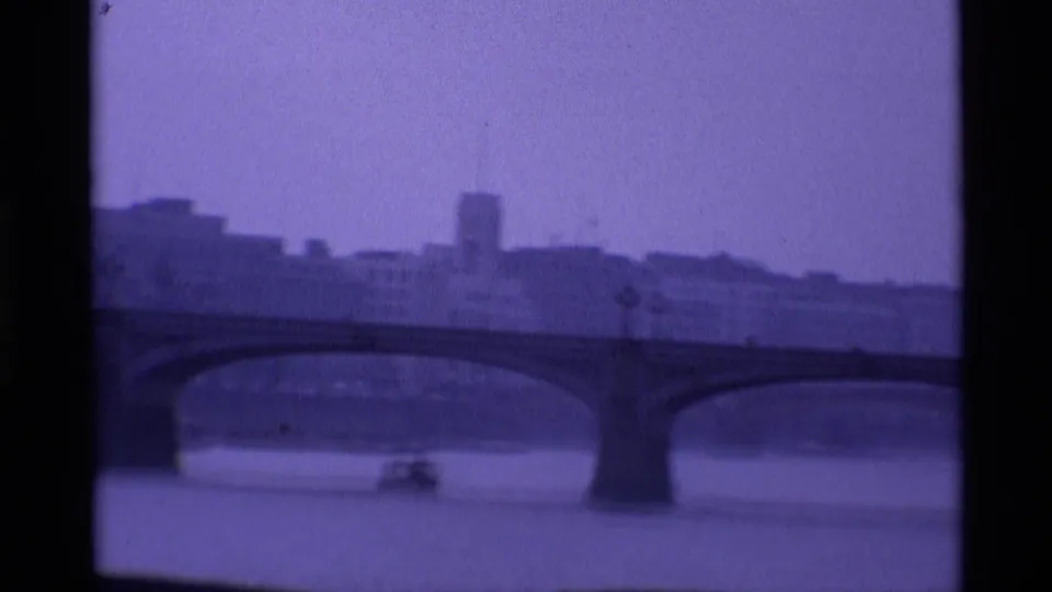 1969 London home-movie frame showing bridge arches across the river and a small boat in the soft, purple-tinted distance.
