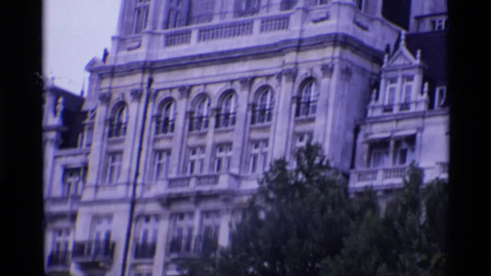 1969 London frame showing a pale multi-story historic facade with arched windows, balconies, dark roof sections, and trees at street level.