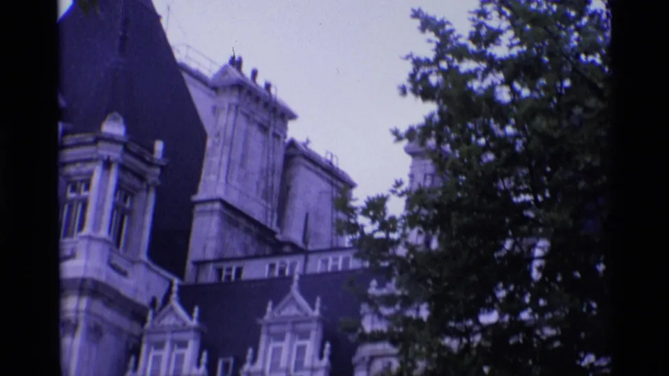 1969 London home-movie frame showing ornate stone buildings with arched windows partly obscured by a leafy tree.
