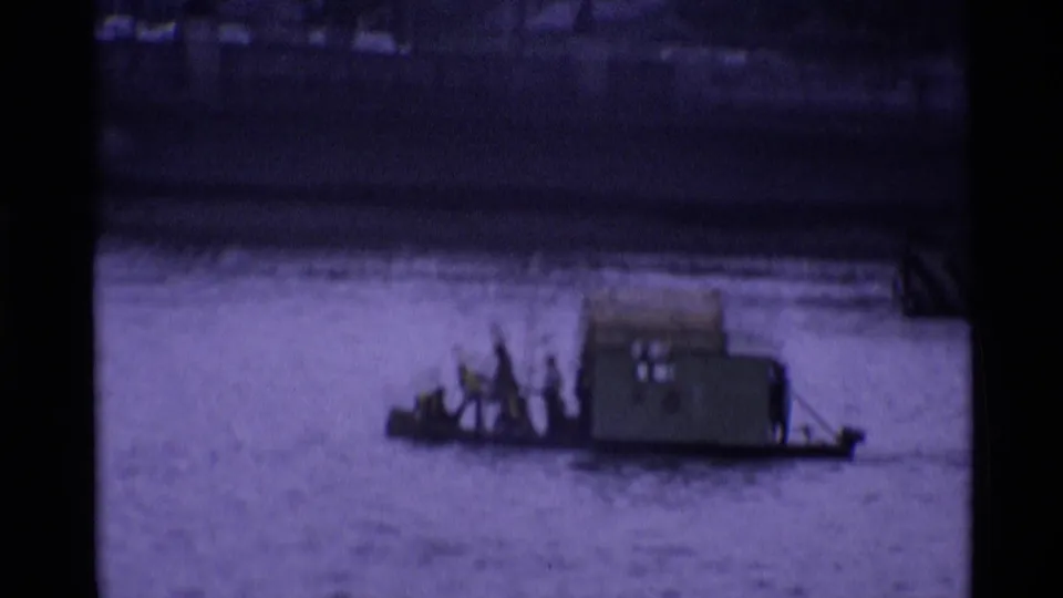 Small river craft with people aboard passes near a stone embankment in 1969 London home-movie footage.